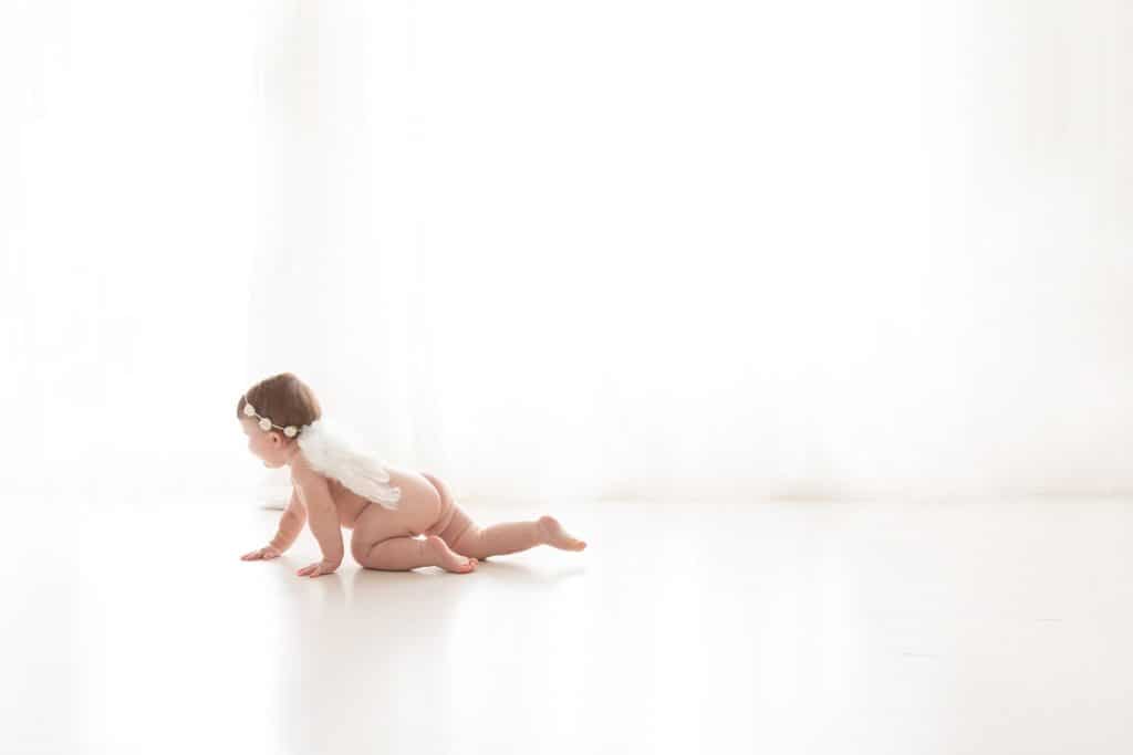 A baby crawls while wearing angel wings. 