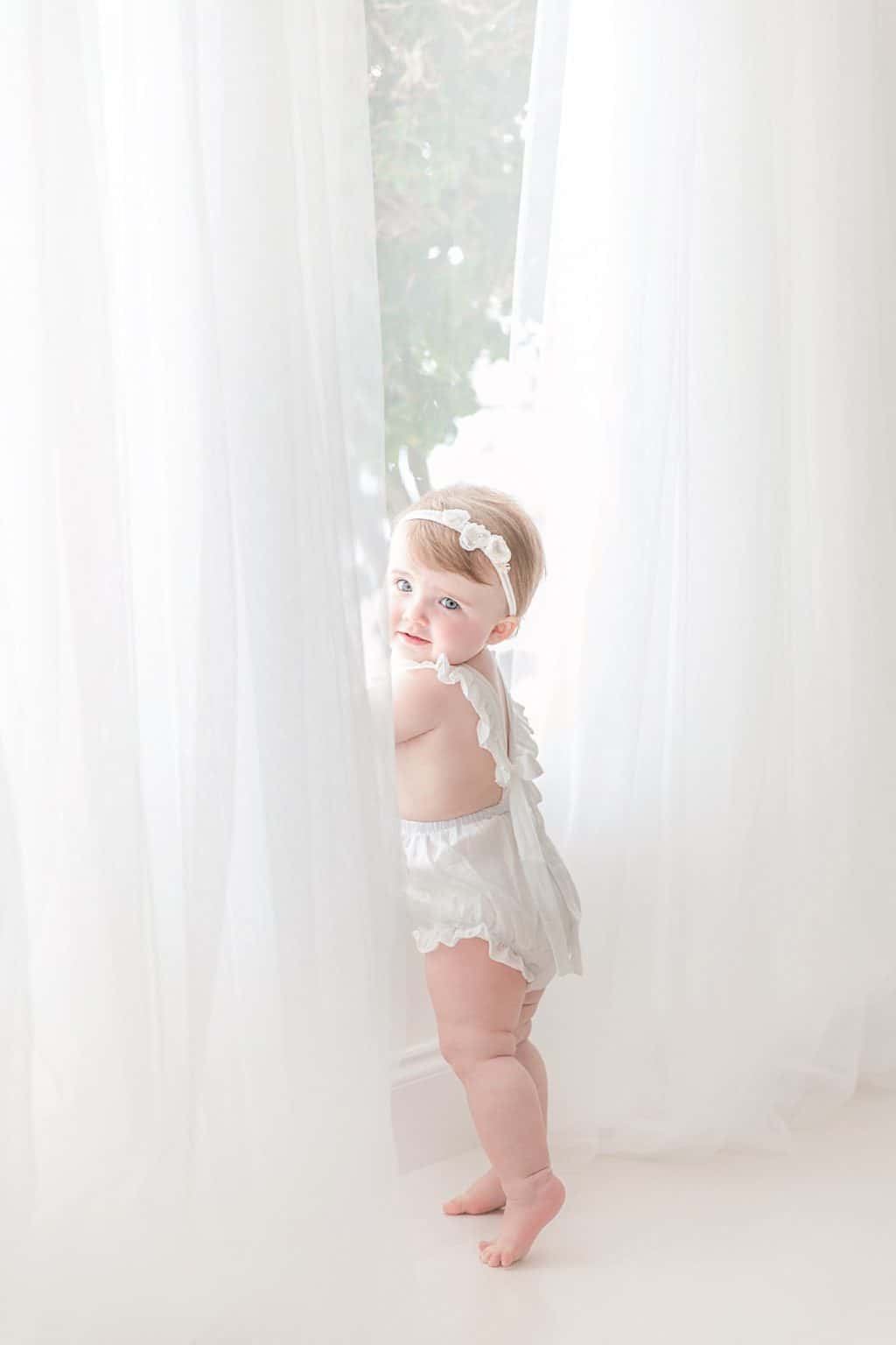 A baby stands by a window with white curtains. 