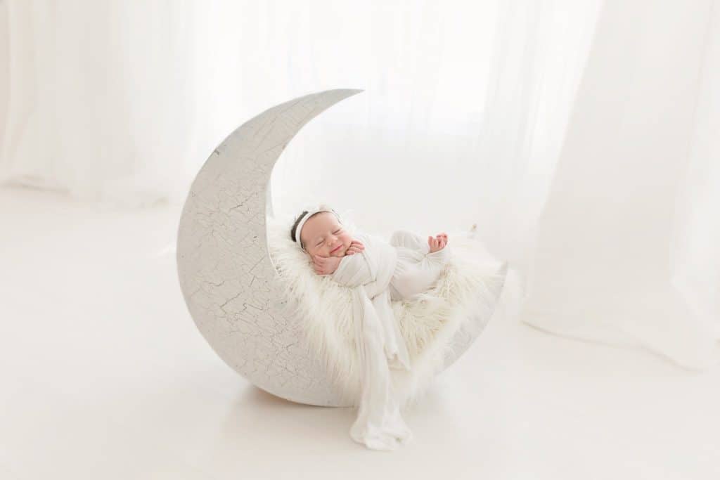 A newborn baby sleeps on a moon shaped photo prop. 