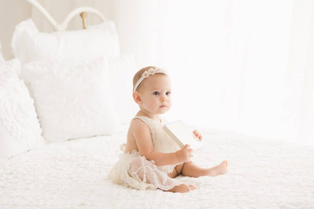 A baby girl sits on a white iron bed with white bedding.