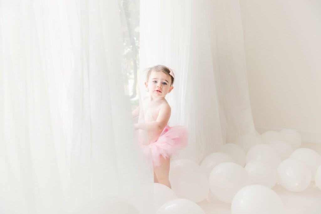 A baby wears a tutu and stands at a window. 