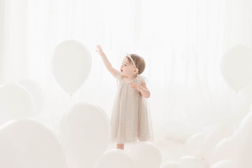 A baby girl reaches up for a white balloon. 
