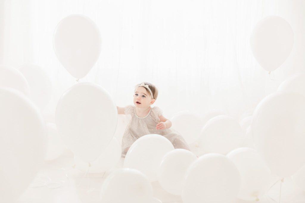 A baby girl sits in a room of white balloons. 