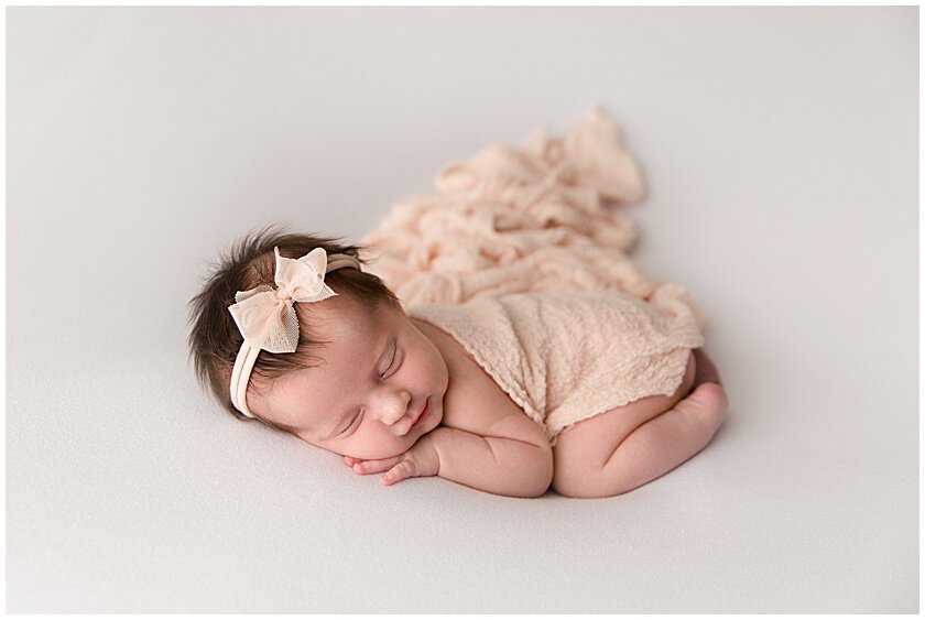 Newborn photos of a baby swaddled in peach with a pink bow while sleeping on a white backdrop in Jupiter Florida photography studio