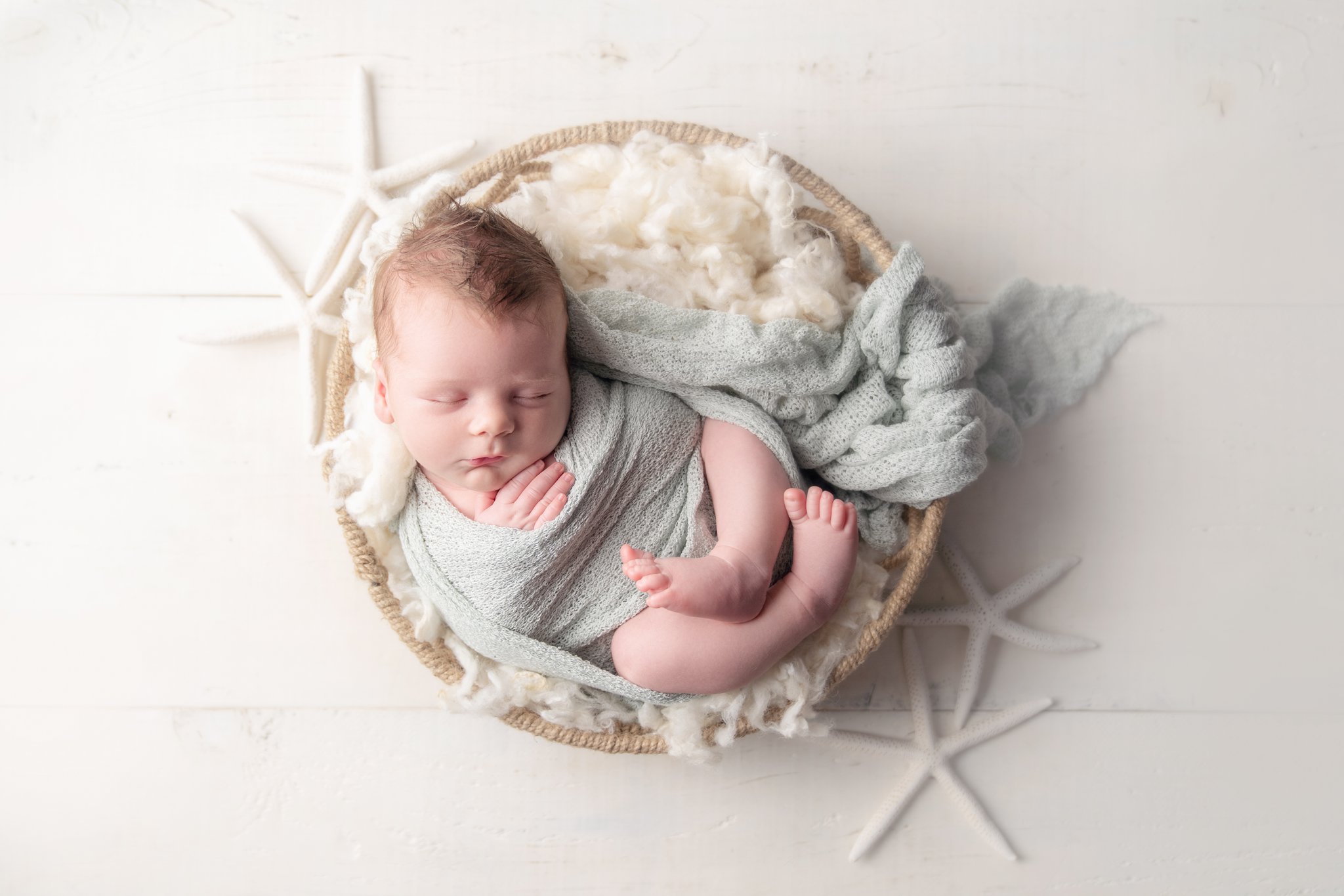 Photo of newborn baby boy being photographed in Jupiter Florida photography studio wrapped in a blue swaddle curled up in a driftwood bowl surrounded by starfish