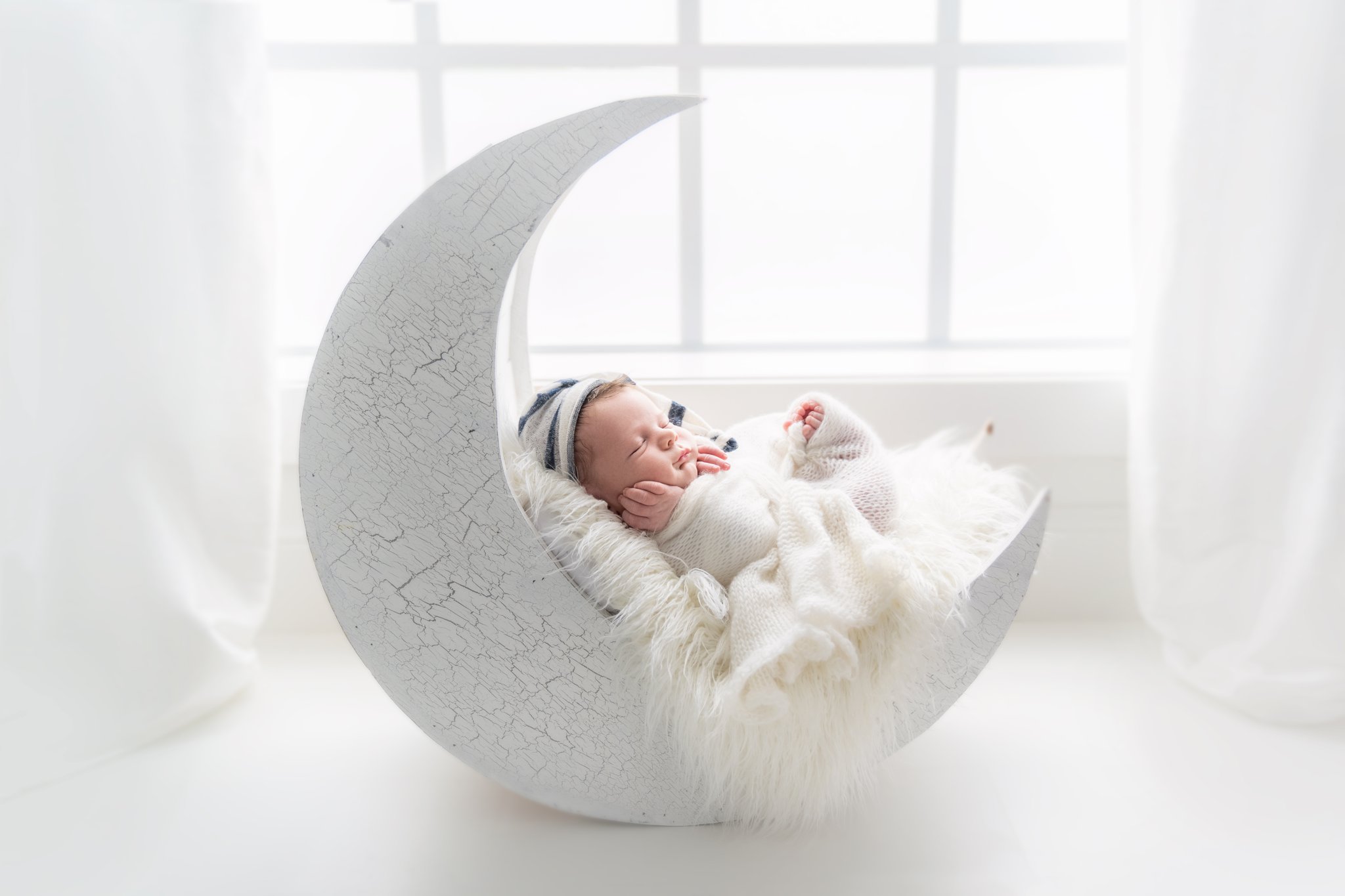 Photo of newborn baby boy being photographed in Jupiter Florida photography studio wrapped in a cream swaddle and navy and cream hat lying on a white moon prop