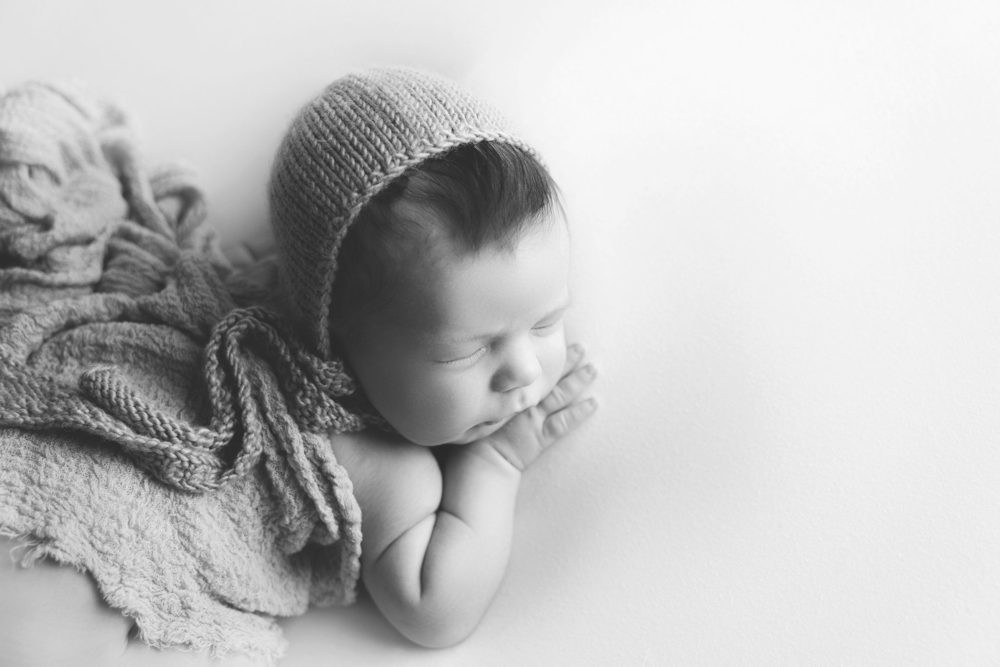 Black and white portrait of newborn baby boy being photographed in Jupiter Florida photography studio on cream backdrop with blue scarf draped over his back and a blue matching knitted bonnet 