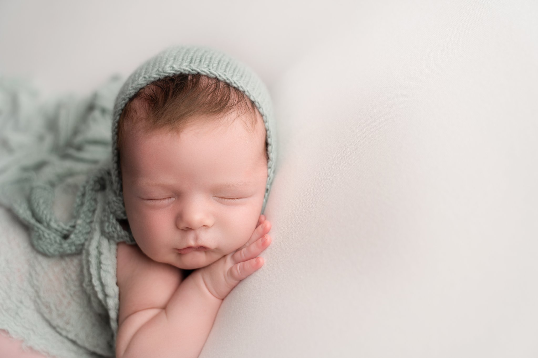 portrait of newborn baby boy being photographed in Jupiter Florida photography studio on cream backdrop with blue scarf draped over his back and a blue matching knitted bonnet 