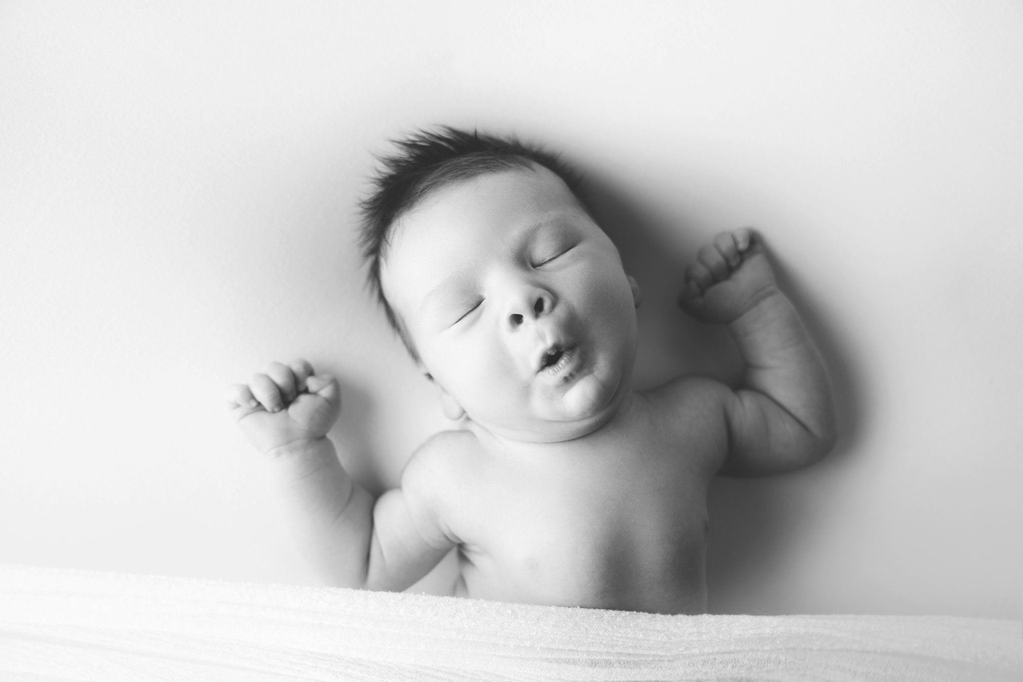 Newborn baby boy being photographed in Jupiter Florida photography studio yawning and stretching while he sleeps on cream backdrop
