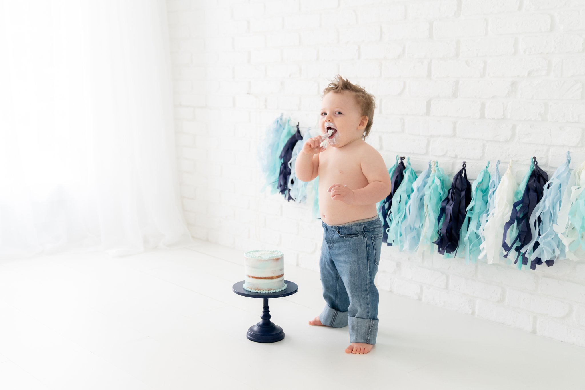 One year old baby in jeans eating and smashing his birthday cake in south florida baby photography studio. 