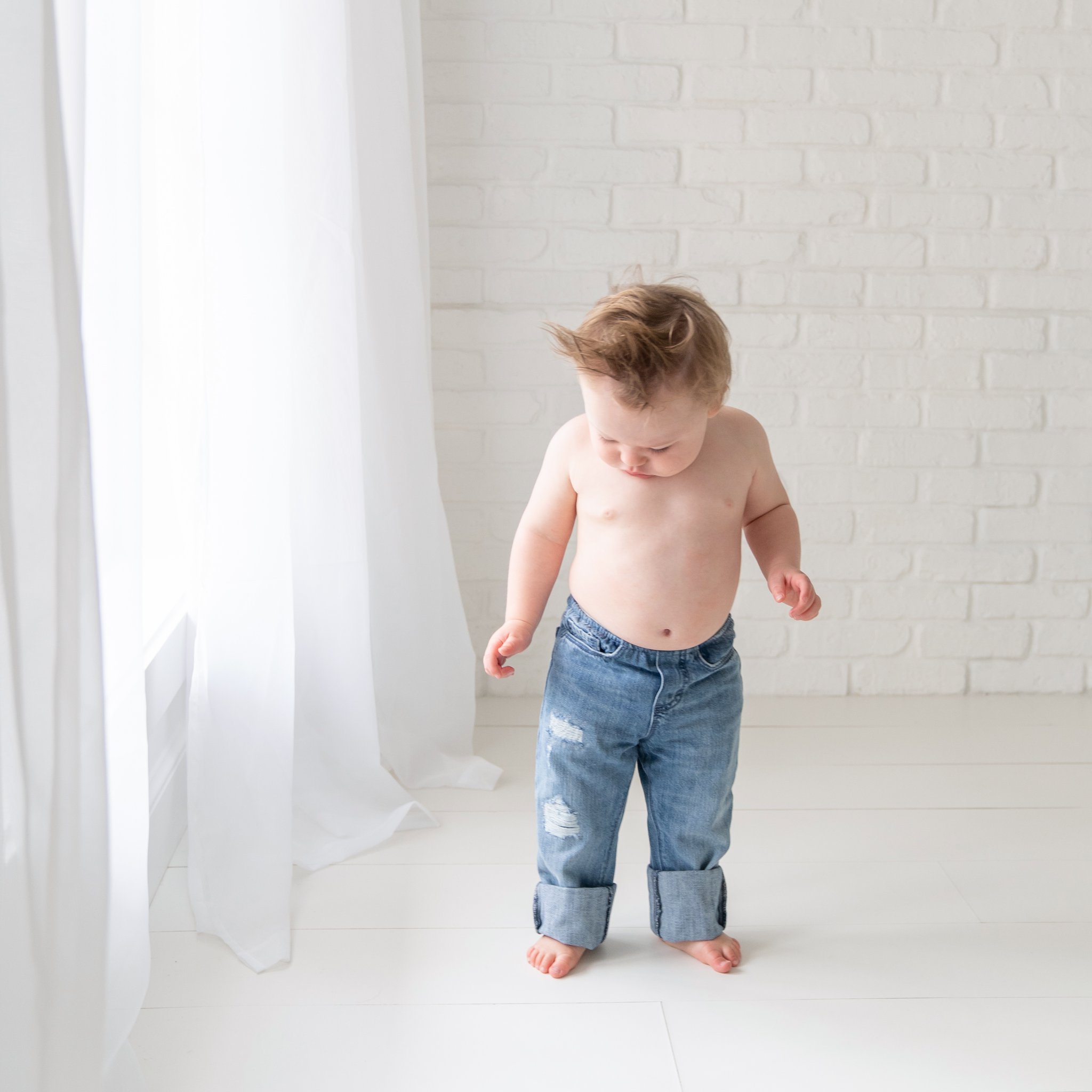 One year old baby in jeans rolled up standing by window in a south florida baby photography studio. 