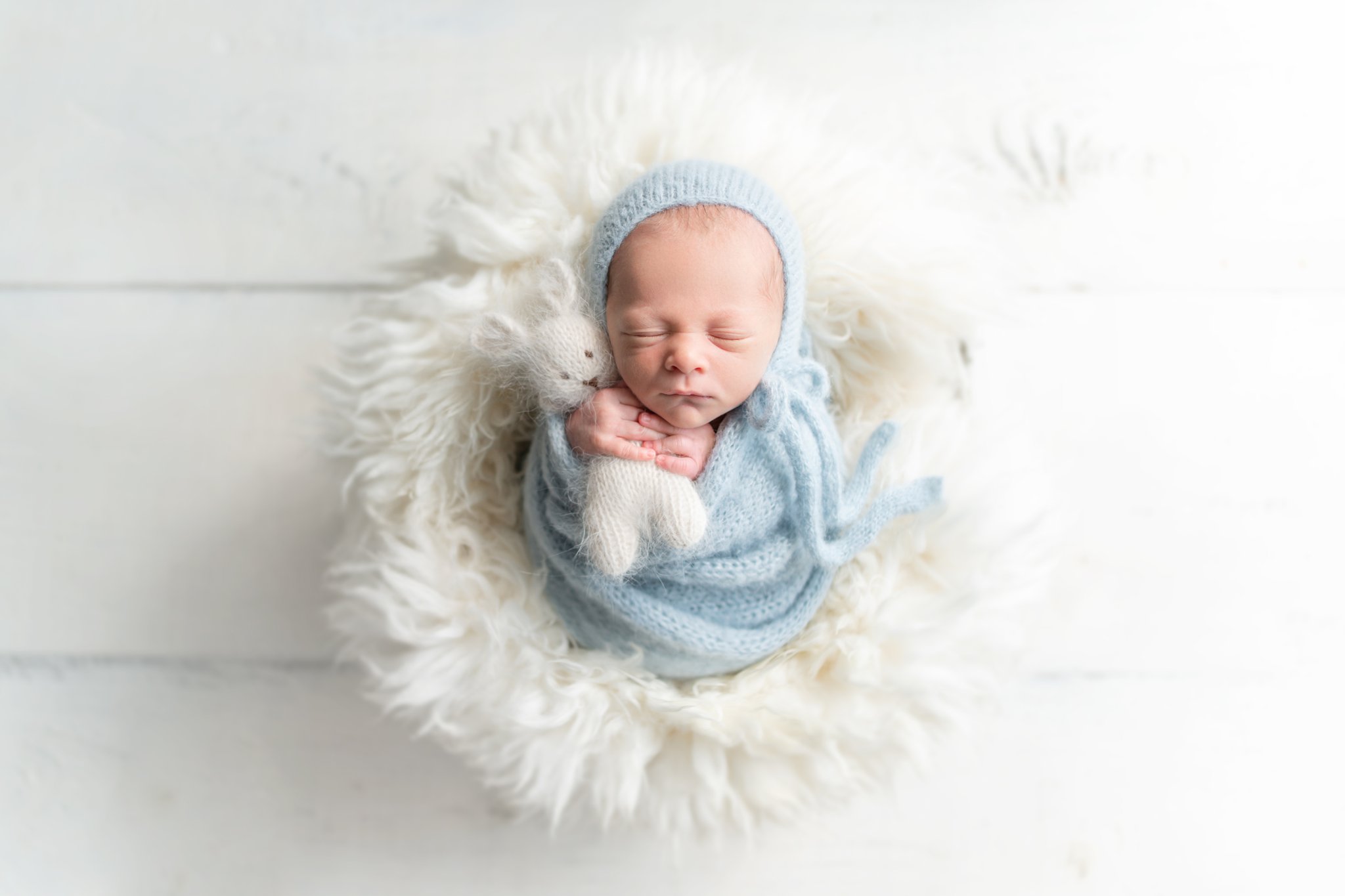 Newborn baby in baby blue swaddle and wearing a blue knitted hat, holding a knitted bunny being photographed in south florida photo studio.