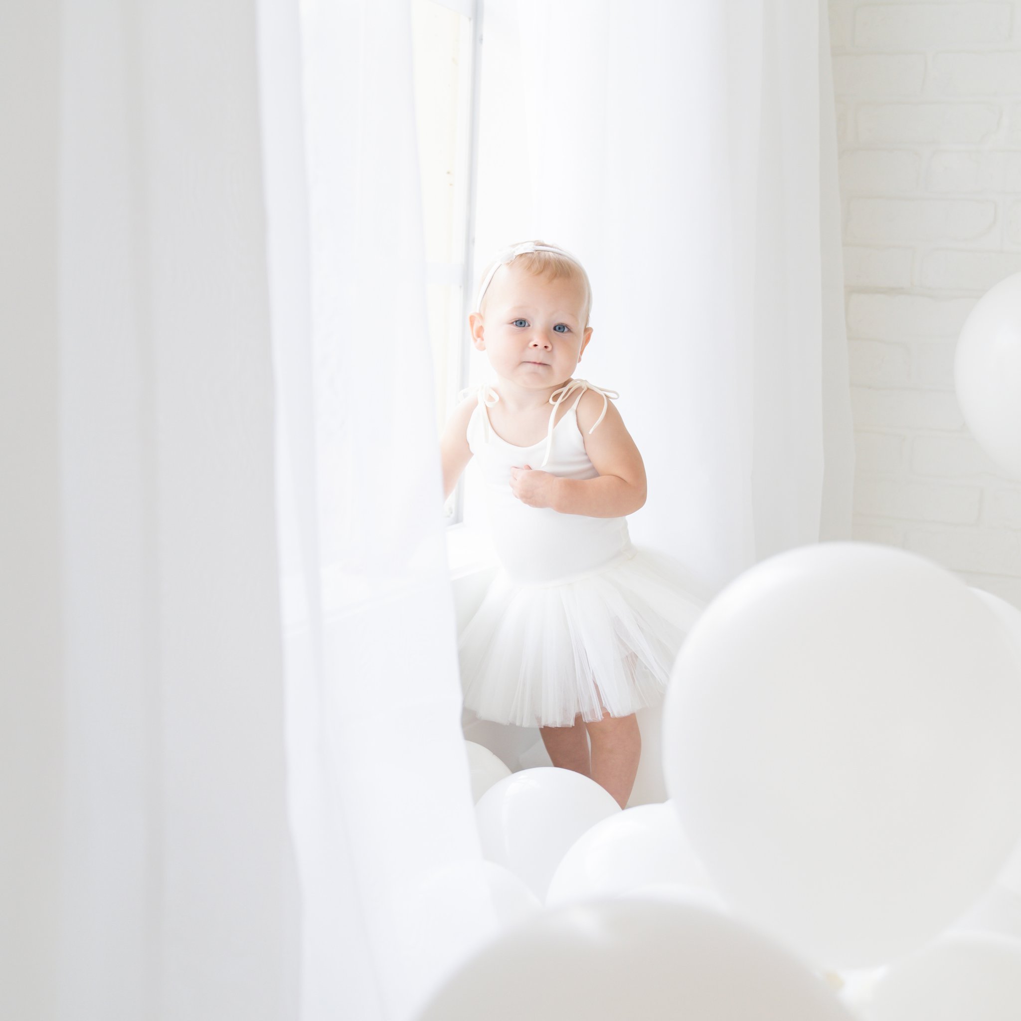 One year old baby, surrounded by white balloons, being photographed next to a large sunny window.