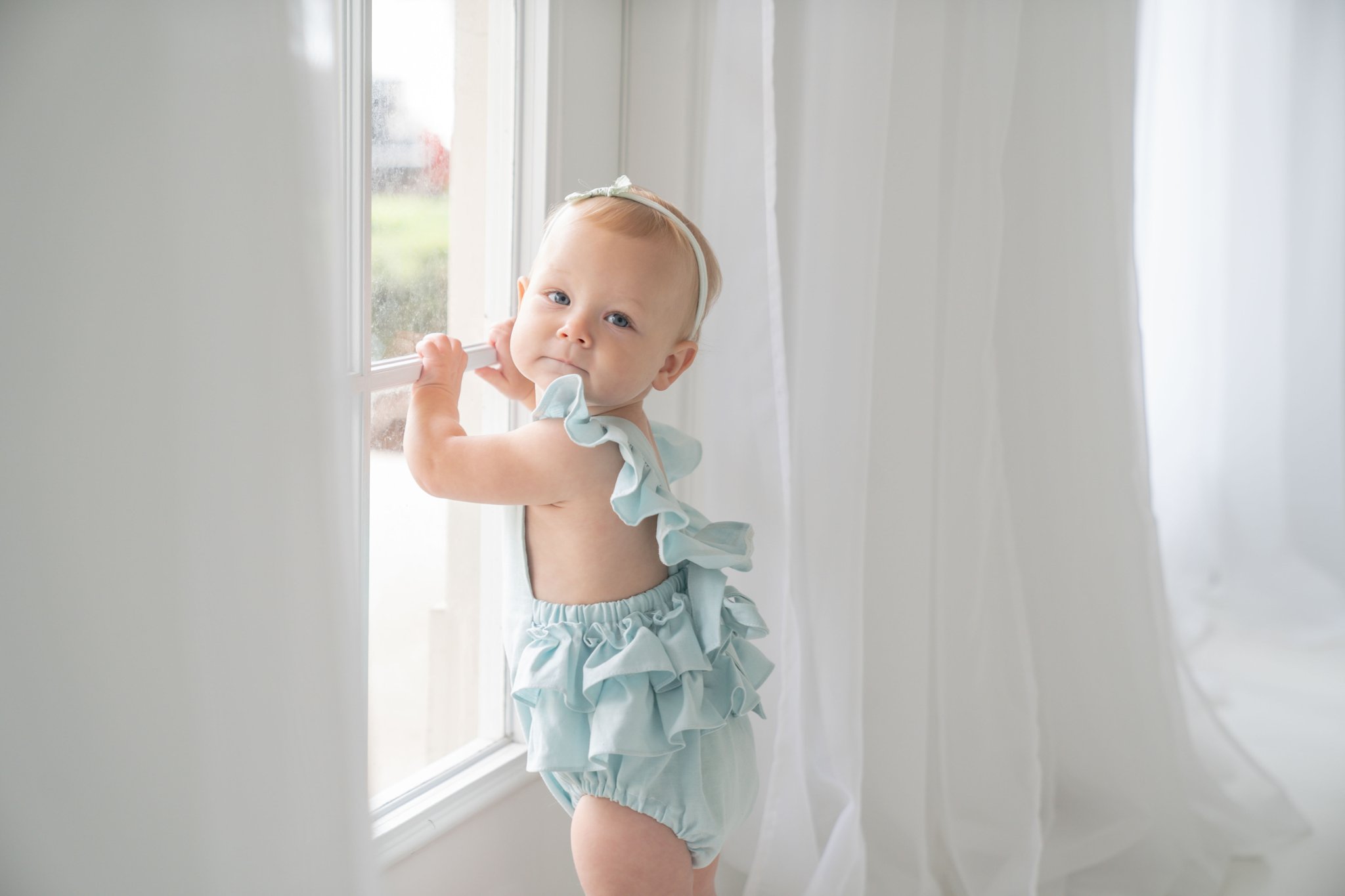 Birthday baby girl being photographed in a south florida photography studio in blue ruffled romper by a white brick wall.