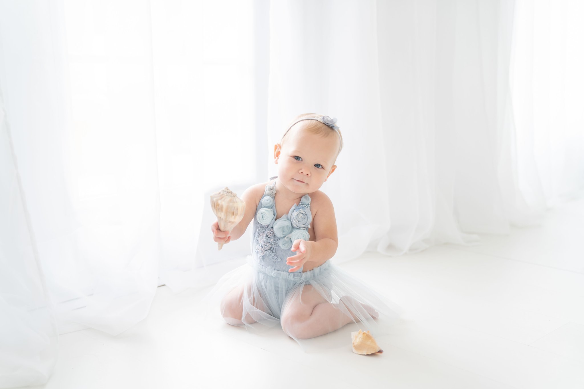 Baby girl being photographed in a south florida photography studio in blue handmade dress while holding beach shells.