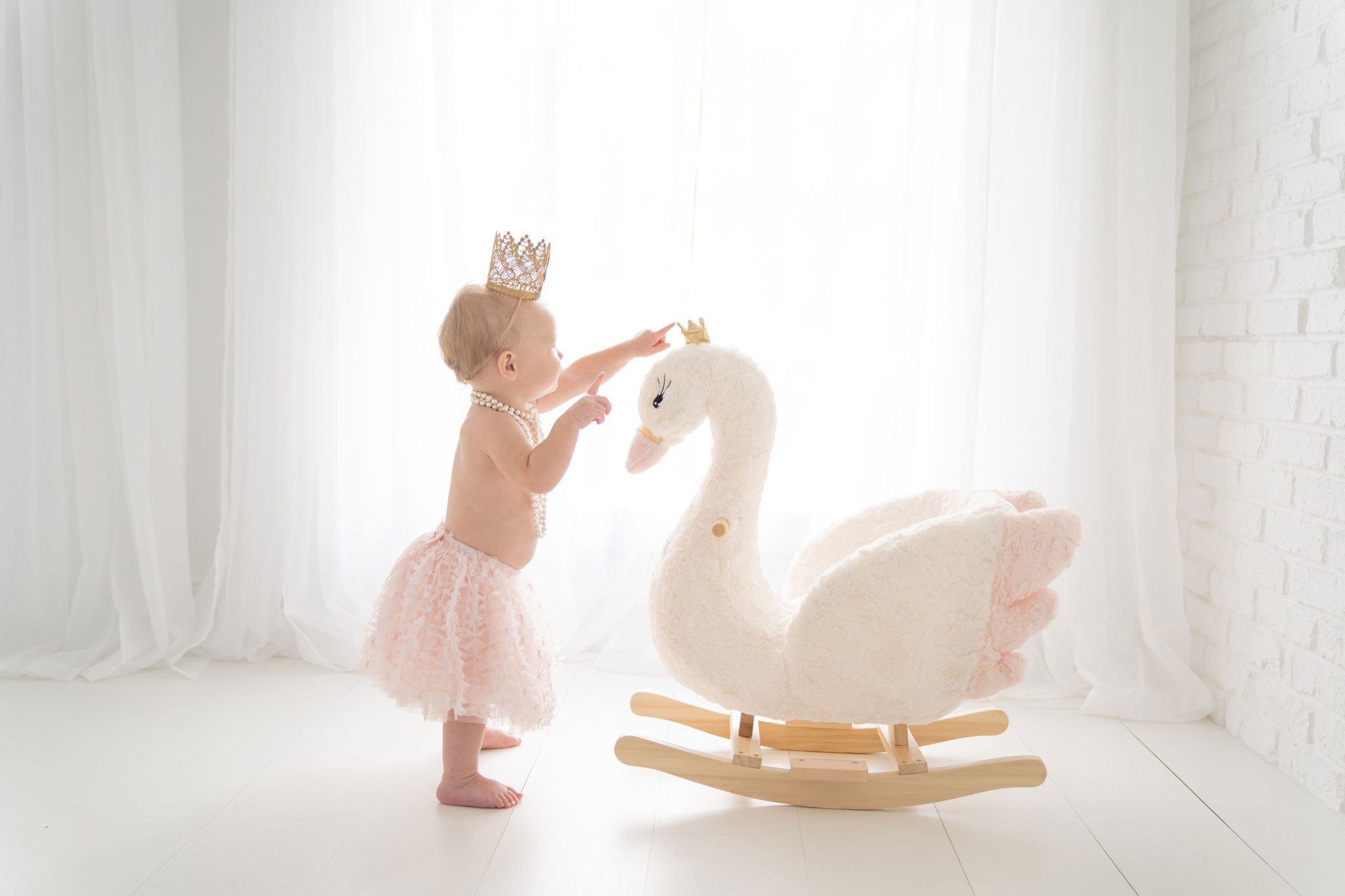 One Year old being photographed in a Jupiter Florida photography studio wearing pink skirt and crown and standing by a rocking swan.