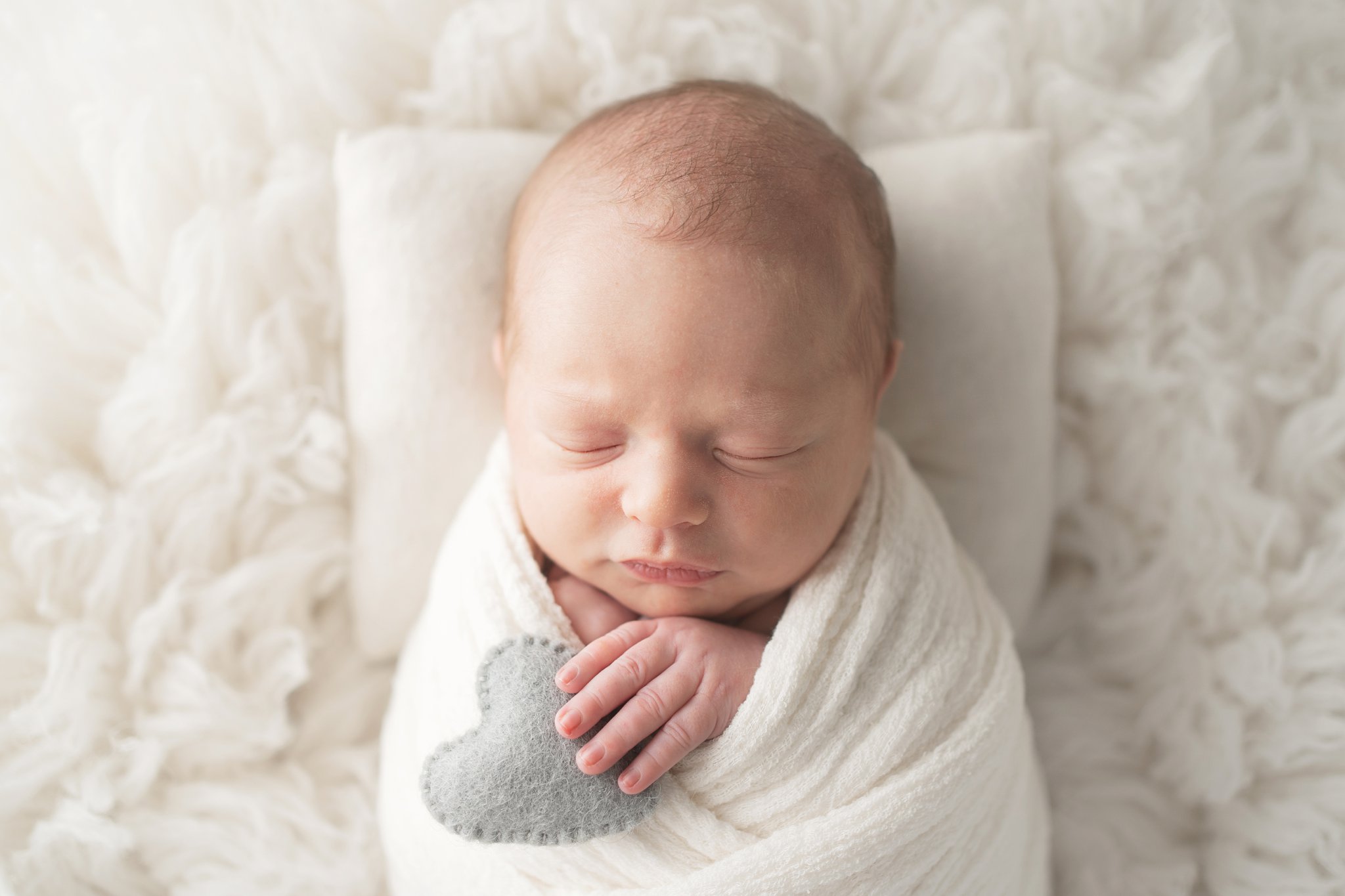 10 day old newborn posing on a white backdrop with a little  grey heart in his hand in jupiter florida photography studio