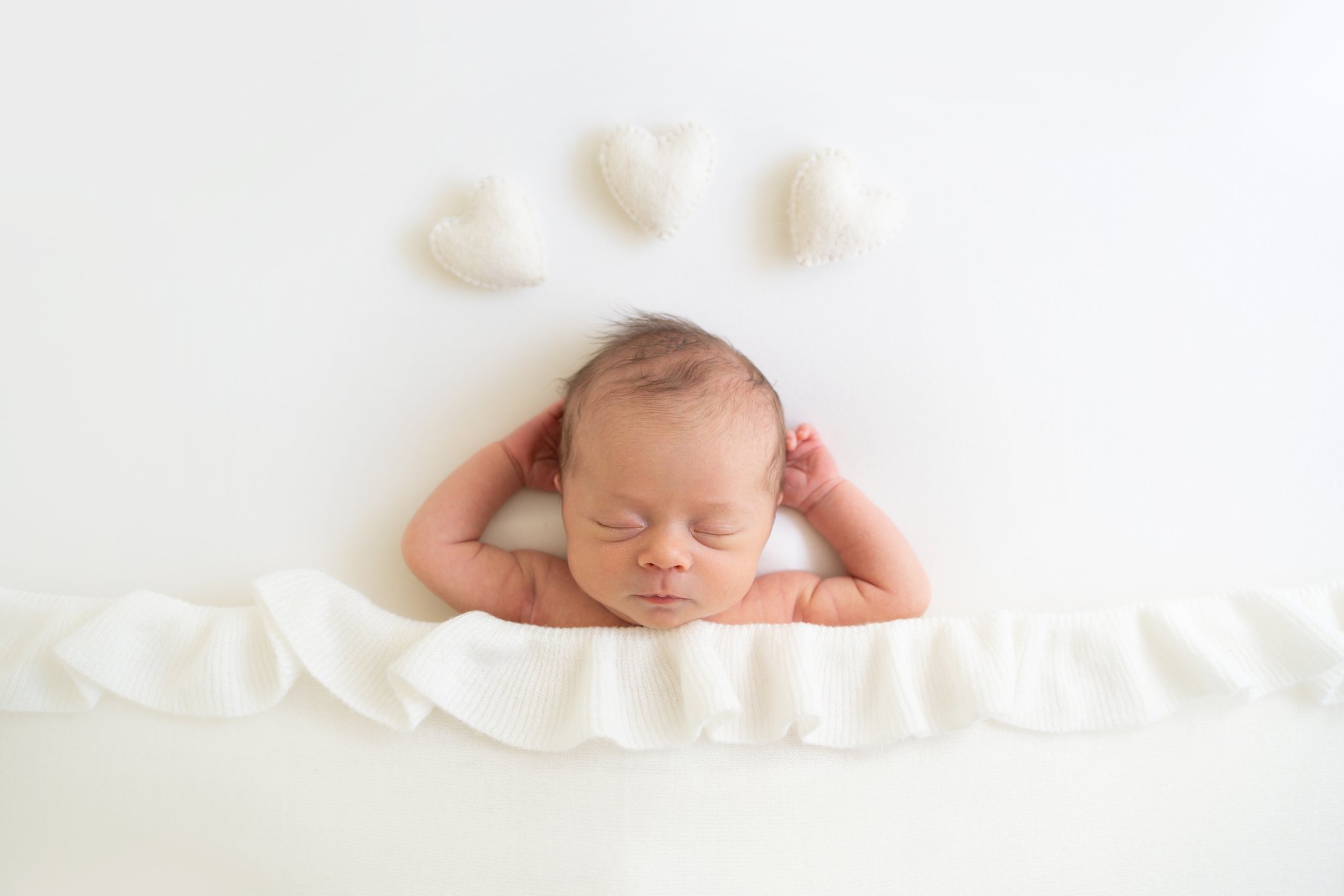 newborn baby laying on bed with white hearts being photographed in jupiter photography studio
