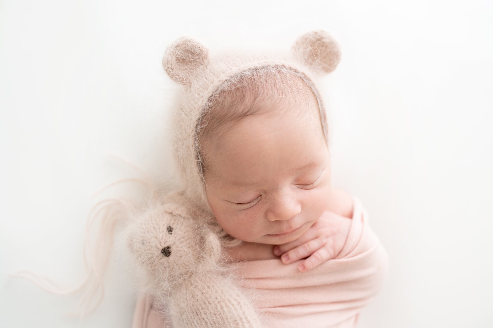 Newborn Photo Shoot featuring 2 week old baby girl wrapped in a pink wrap with a pink bear hat.