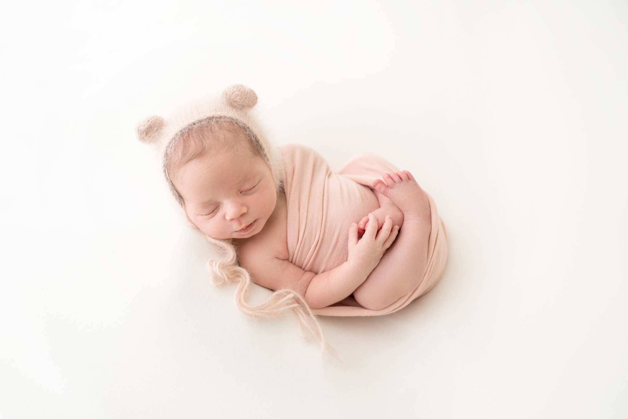 Newborn Photo Shoot featuring 2 week old baby girl wrapped in a pink wrap with a pink bear hat.