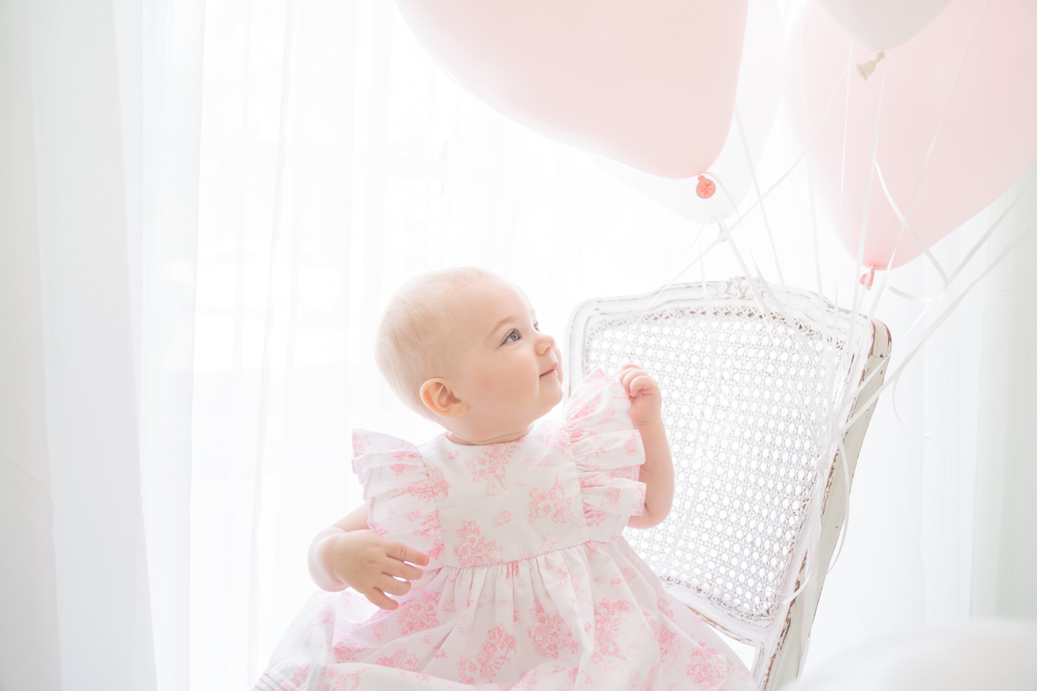 Birthday baby with pink and white toille dress sitting on a vintage chair in a room full of balloons