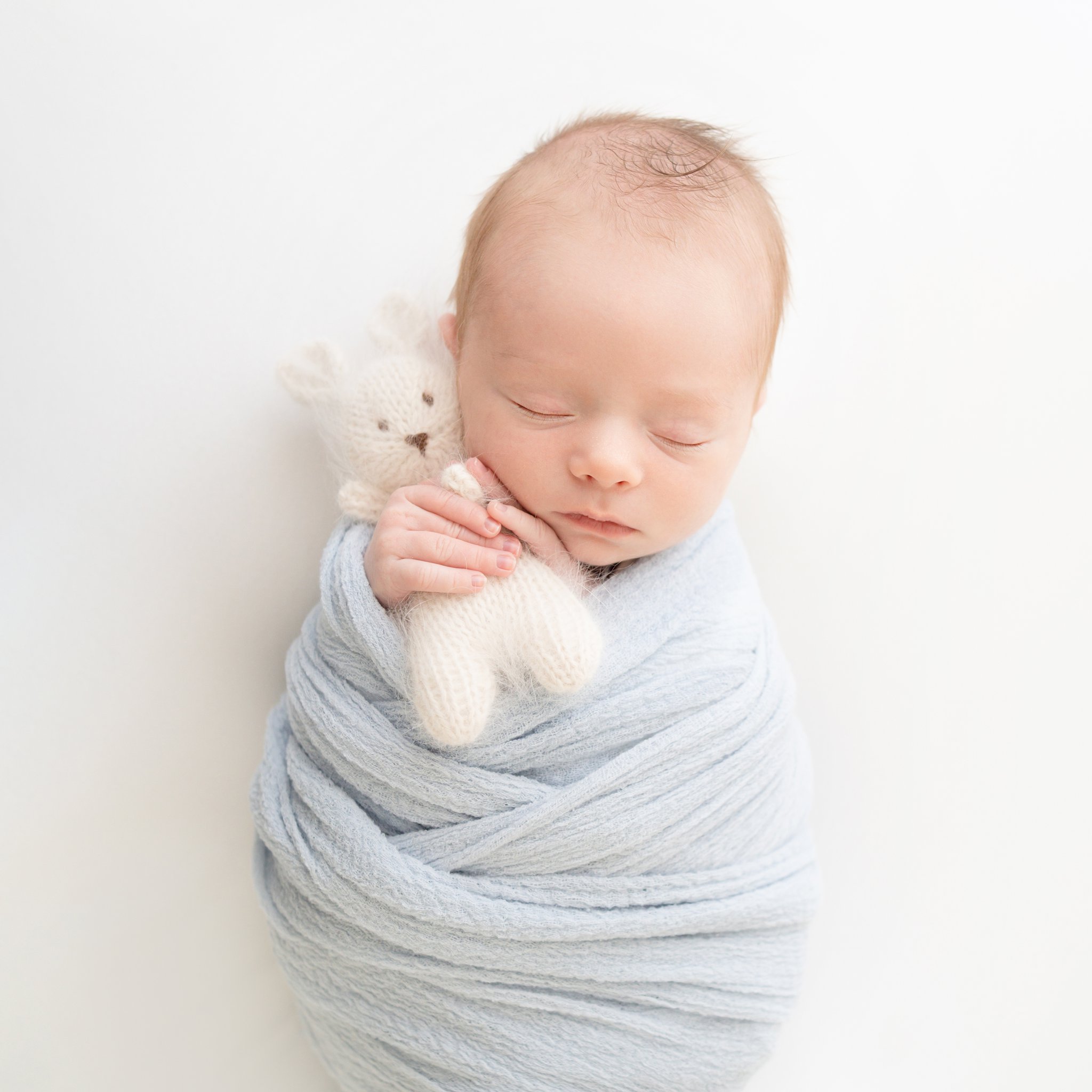 Newborn swaddled in blue holding soft white knit bunny stuffed animal. 