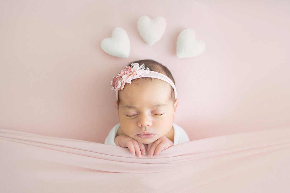 Newborn baby girl swaddled in a pink wrap lying on her back on a pink backdrop with hearts over her head