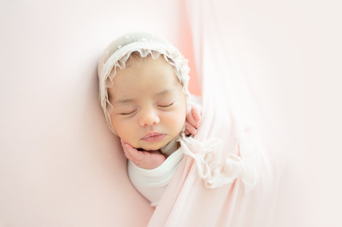 Newborn baby girl swaddled in a pink wrap lying on her back on a pink backdrop with a bonnet on her head