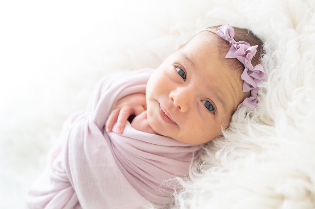 Newborn baby girl laying on her back with a purple bow headband on her head while she looks at the camera