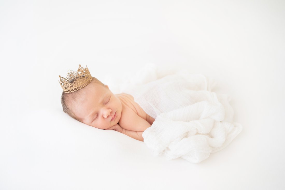 Newborn baby lying on her tummy with white wrap covering her wearing a princess crown on her head.