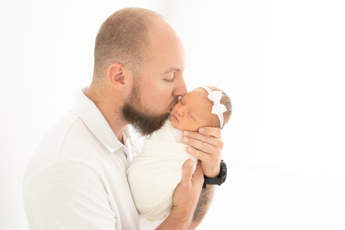 Newborn baby swaddled in a cream wrap being kissed on the cheek by her dad