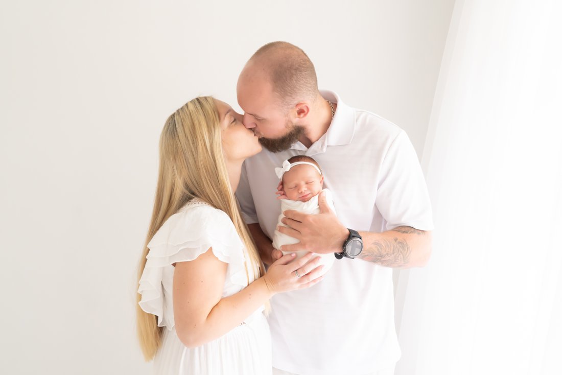 New mom and dad kissing holding their smiling newborn baby while being photographed.