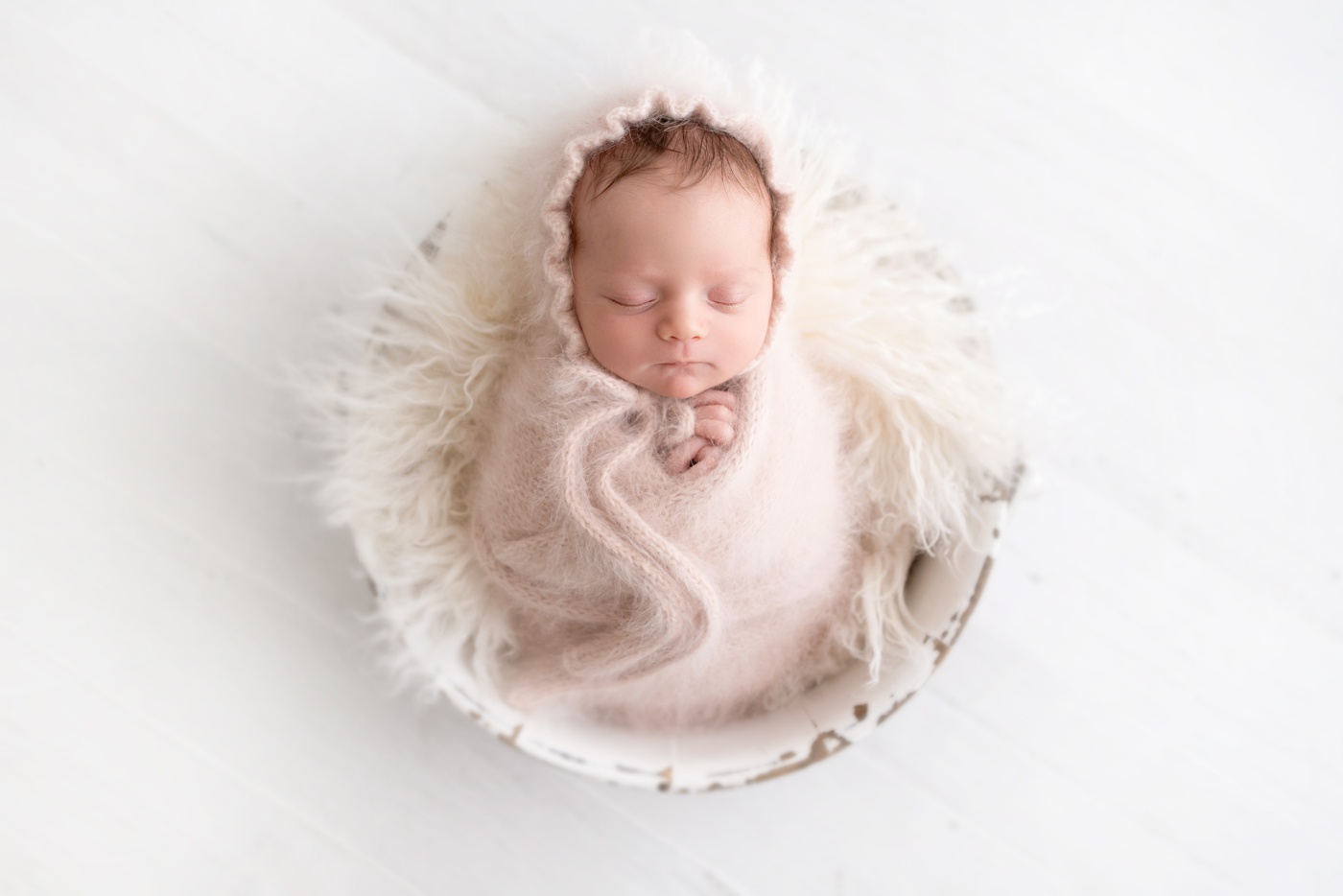  newborn baby girl wrapped in pink angora wrap and bonnet laying in a white antique bucket