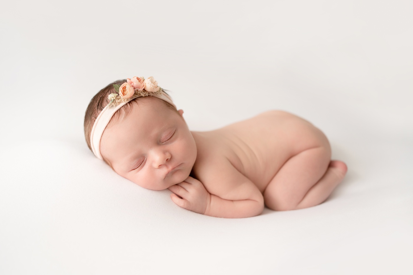  newborn baby girl laying on cream backdrop woith a pink headband sleeping curled up with her hand under her chin