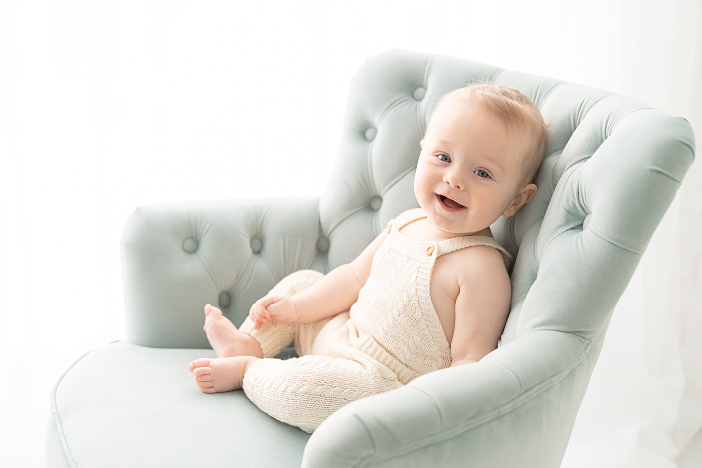 baby boy wearing white overalls laughing in a turquoise velvet chair
