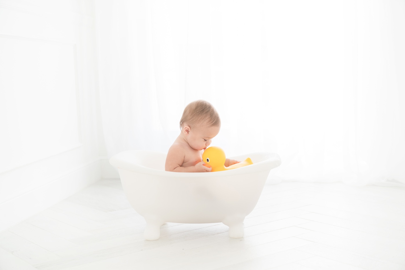 Baby girl in a in tiny white bathtub by a natural light window being photographed for her one year photoshoot