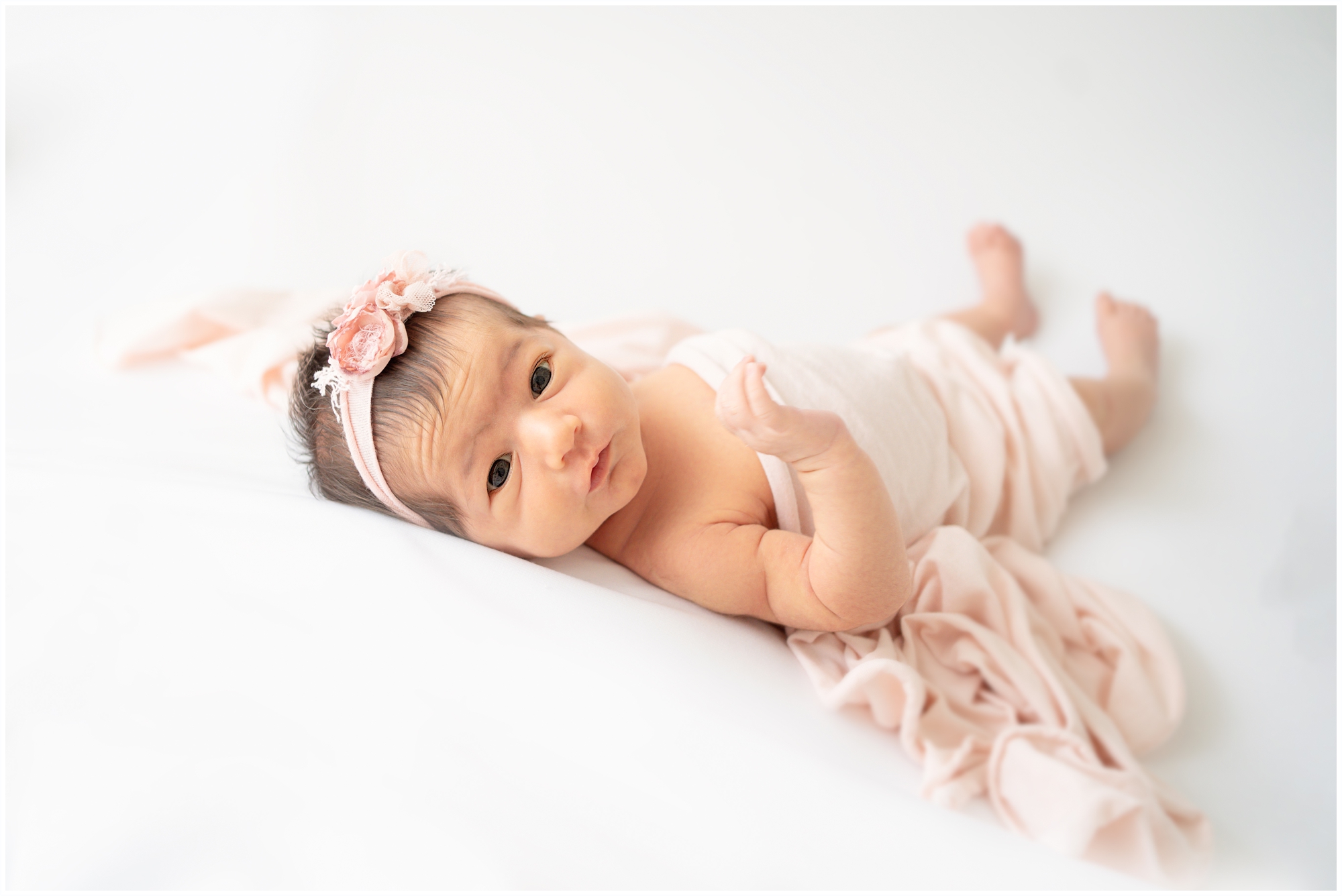 Newborn photoshoots of a new baby girl wide awake looking into the camera while laying on her back swaddled in a pink wrap