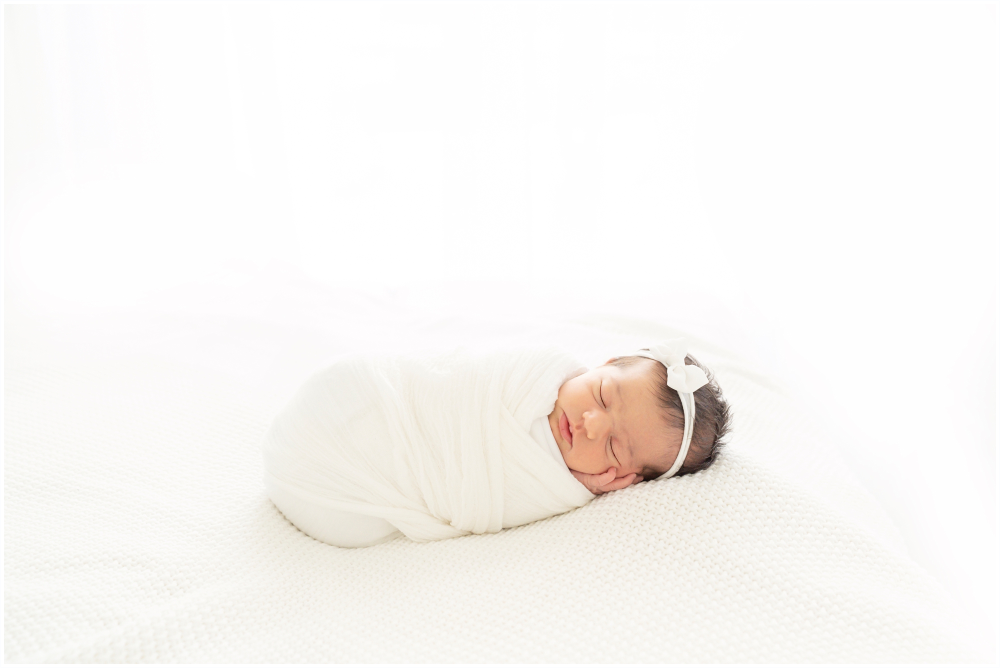 Brand new baby swaddled in white laying on a white bed being photographed in Jupiter Fl studio