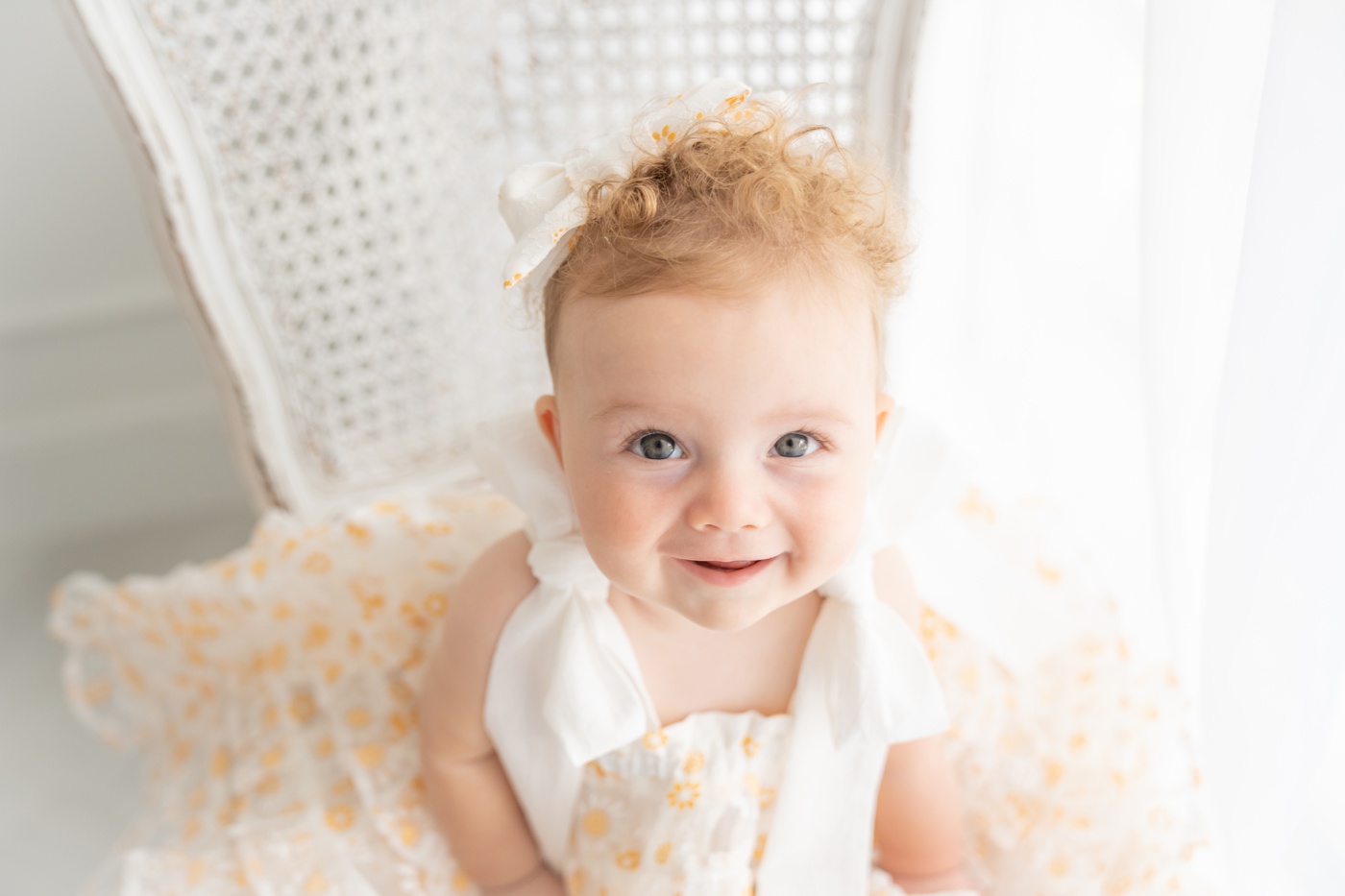 Baby girl sitting on an antique white chair in a white fluffy dress with yellow daisies