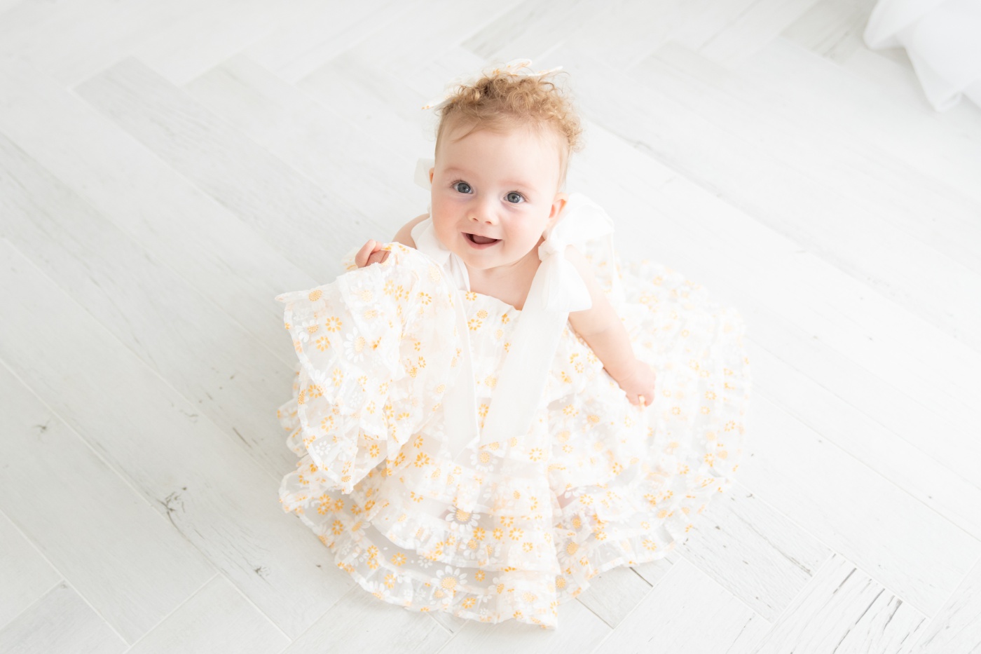 Baby girl sitting the floor of a white photography studio wearing a white fluffy dress with yellow daisies