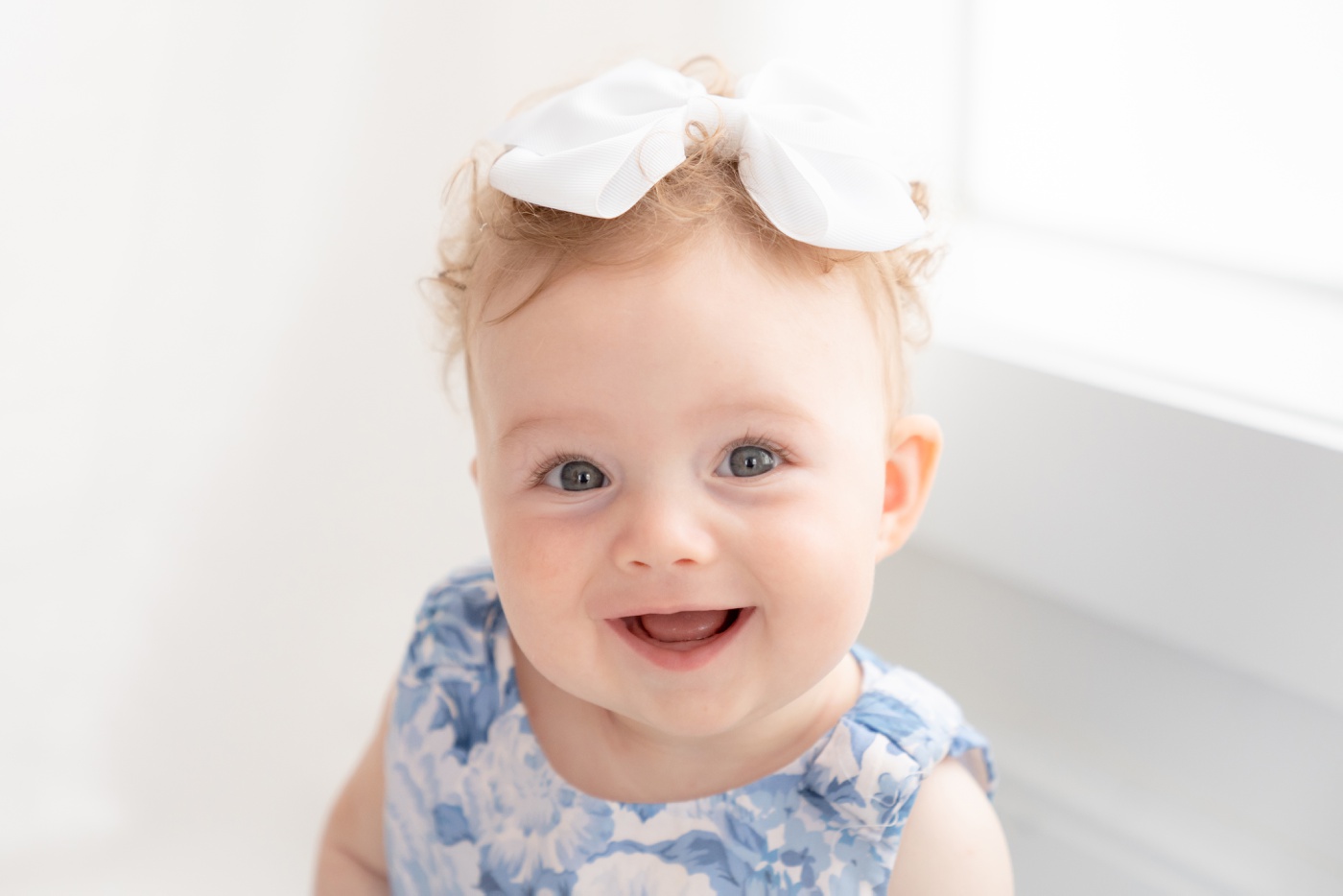 Baby girl sitting the floor of a white photography studio wearing a white and blue floral dress
